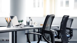 Aparatos modernos en el interior de una oficina de coworking durante la pandemia de COVID-19. Sillas y escritorio para trabajadores de apoyo, computadoras portátiles y auriculares, tazas y artículos de papelería a la luz del día.